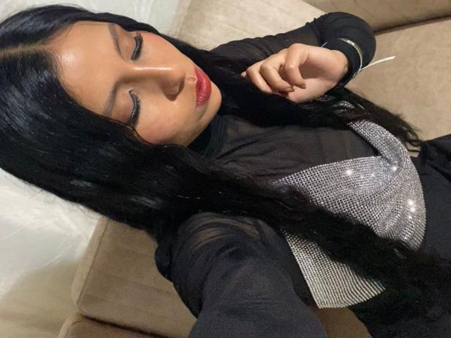 IsabelEvanss with long black hair, red lipstick, closed eyes, wearing a black top with silver sparkly fabric, taking a selfie, holding her chin, on a beige couch.
