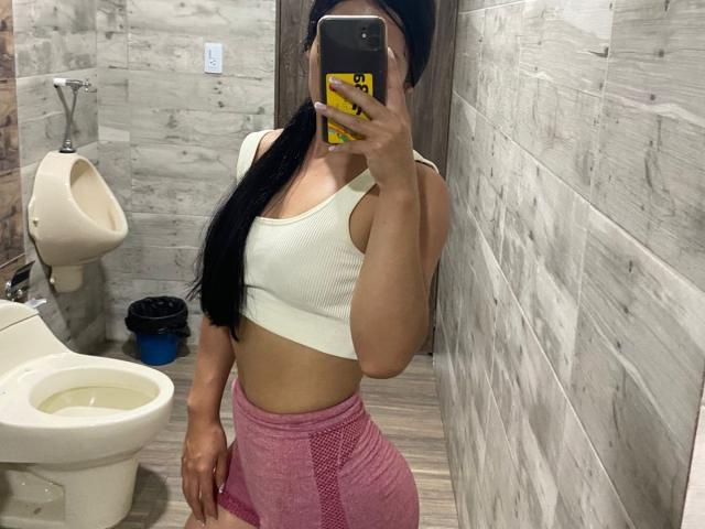 IsabelEvanss in a bathroom mirror selfie, wearing a white sports bra, pink mesh gym shorts, black beanie, holding a yellow phone, with a visible urinal and toilet.
