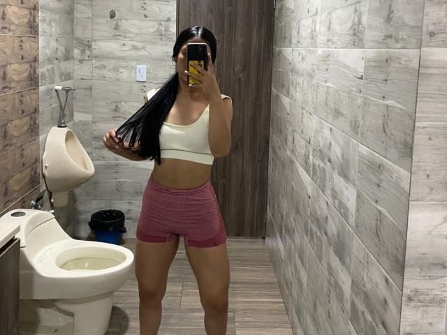 IsabelEvanss in a bathroom, wearing a white sports bra and pink shorts, holding a phone with a yellow case, showing her long black hair and tan skin.