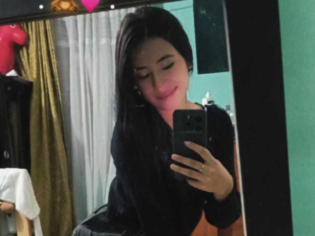 Helen Watson, con lunghi capelli neri, sorridente, indossa una maglietta nera, tiene in mano un telefono, sta scattando un selfie allo specchio, mostrando leggermente il seno, in una stanza con pareti verdi..