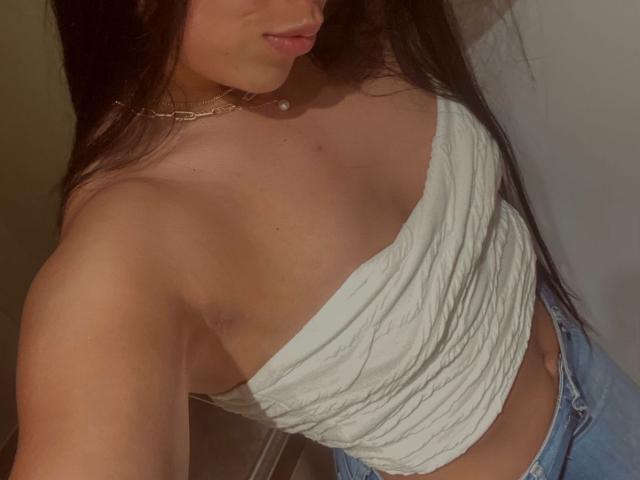 IsabelEvanss with long brown hair, wearing a white, ruched, one-shoulder top that shows her tits, and blue jeans, showing her tan, smooth skin and a delicate necklace.