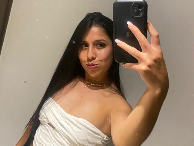 IsabelEvanss with long black hair, wearing a white strapless top, holding a black phone, taking a mirror selfie, showing her tits, with a subtle smile and pink lips.