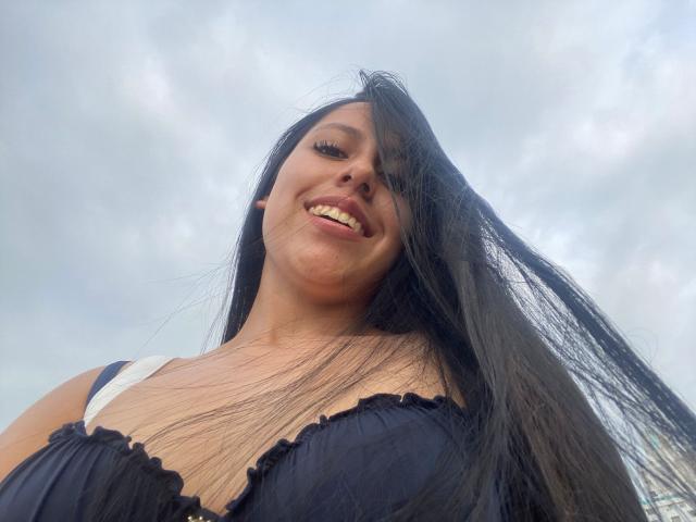 IsabelEvanss with long black hair, smiling down at the camera, wearing a black top with a white strap, her tits slightly visible, under a cloudy sky.