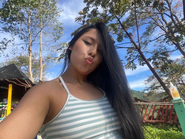 IsabelEvanss with long black hair, pouty lips, wearing a striped tank top, outdoor shot, blue sky, green trees, red wooden fence, yellow and green building in background.
