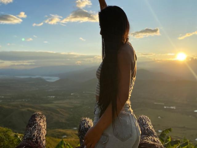 IsabelEvanss with long dark hair, raising arm, wearing striped tank top and denim shorts, sitting on wooden fence, sunlight, cloudy sky, green landscape, sunset, back to camera.