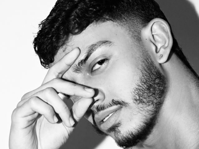 AxelStein with curly hair, medium beard, and earring, covering his eye with his finger, showing a subtle smirk in a black-and-white photo. His nose and lips are visible.