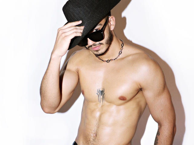 AxelStein, shirtless, wearing a black hat and sunglasses, has a chain necklace, a spider tattoo on his chest, and a trimmed beard, with a muscular, tan, and hairy body.
