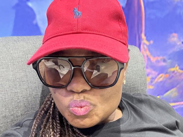 CuteBri with red cap, black sunglasses, and pink pout, puffy lips, wearing black shirt, and braided hair, looking straight at camera. Boob-like reflection in sunglasses.