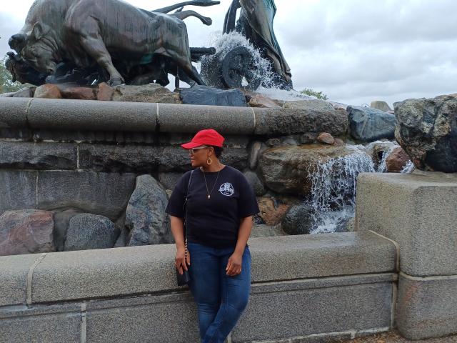CuteBri stands in front of a statue, wearing a red cap, black shirt, and blue jeans, with a waterfall and rocks behind her. Her tits are small, and she's cute.