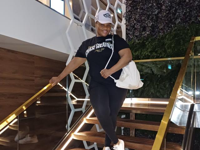 CuteBri, dark-skinned woman with a curvy ass, wearing a black "CHAMP BREATHE" shirt, black leggings, white cap, white bag, and white shoes, climbing wooden stairs.