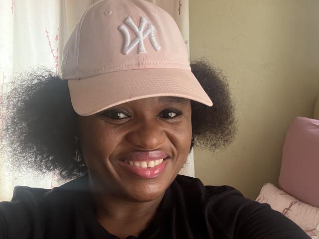 CuteBri has dark skin, curly black hair, and is smiling with pink lipstick. She's wearing a pink NY baseball cap and a black shirt.