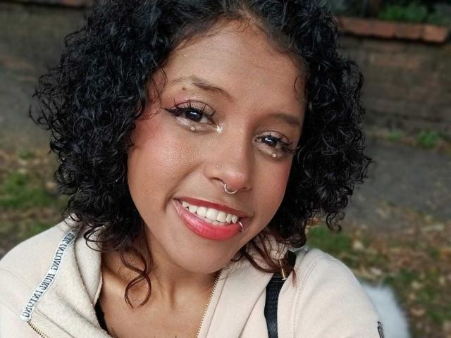 LunaKeller with curly black hair, brown skin, red lipstick, nose ring, eyeliner, and black lip ring, smiling with tears on her cheek. She's wearing a beige hoodie.