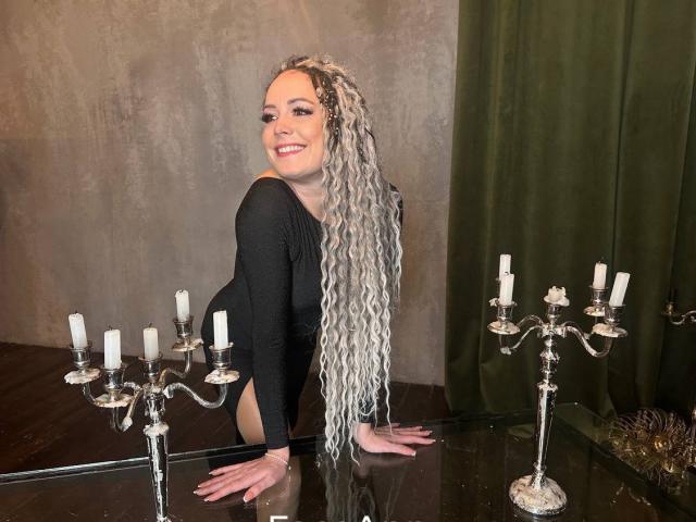 SamanthaReider with long gray-blue braids, black long-sleeve bodysuit, smiling, crouched on glass table, two silver candelabras with white candles, in dimly lit room.