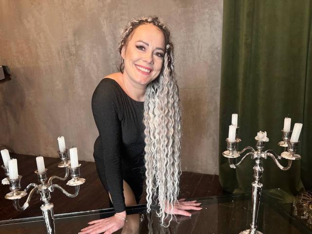 SamanthaReider leans forward on a glass table, smiling at the camera. She has long, wavy silver hair and is wearing a black long-sleeve top. Her hands rest on the table, which has a silver candelabrum with white candles on both sides. The candelabrum is positioned on the left and right, creating a balanced composition. The background is a textured, beige wall with a green curtain on the right. The image is brightly lit, highlighting her smile and hair.