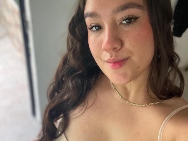 GagingZhara with wavy brown hair, light pink lips, and a subtle smile, wearing a thin gold chain necklace, and a beige strap top, showing her cute, small tits.