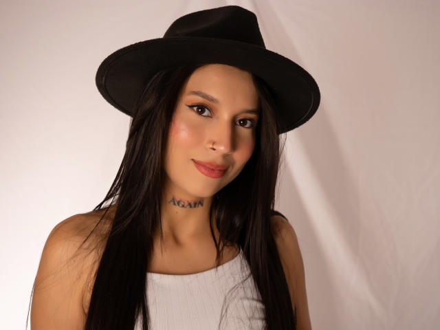 CloeThonsom with long black hair, wearing a black hat and white top, has a tattoo under her chin, and a subtle smile, with brown eyes and pink lips.