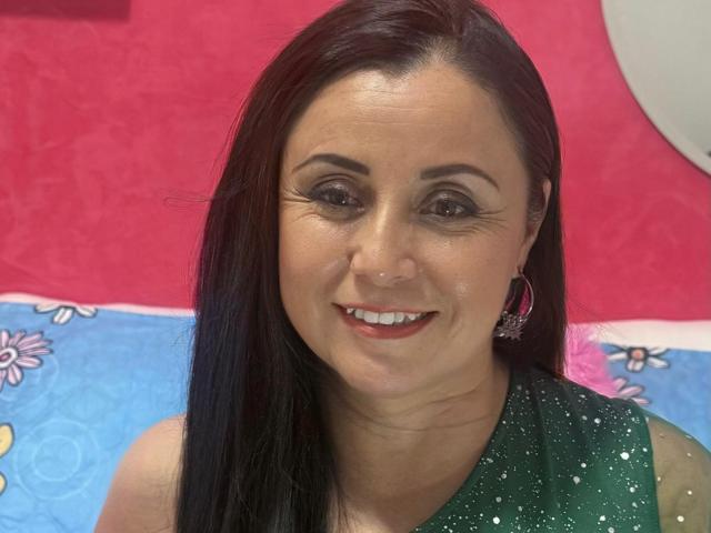 AmandaCooperX has dark hair, brown eyes, and a shiny smile with red lipstick. She's wearing a green, sparkly top and pink earrings. Her ass is not visible.