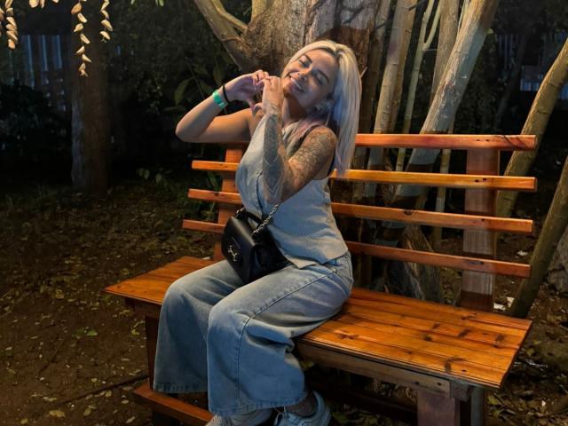 MajoJones with bleached blonde hair, tattoos on her right arm, wearing a blue denim overall dress, black crossbody bag, sitting on a wooden bench, night, outdoors, touching her chin.