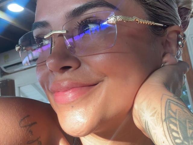 MajoJones with glasses, smiling, holding cheek, tattoo on arm, sunlit skin, small nose, pink lips, gold-tinted frames, blue light reflection, short brown hair, relaxed expression.