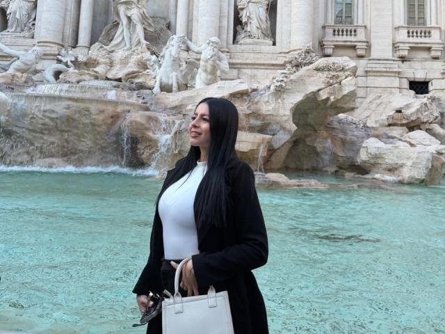 IsabellaJames with long black hair, white top, black coat, holding white bag, standing by turquoise water, smiling, rocks and water feature in background.