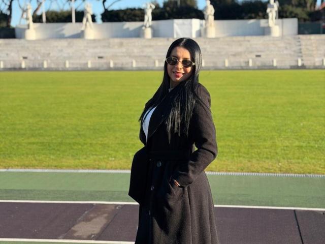 Isabella James, con cabello negro largo, vestida con un abrigo negro, una blusa blanca y gafas de sol, con las manos en los bolsillos del abrigo, de pie en un campo de hierba verde, mostrando su trasero..