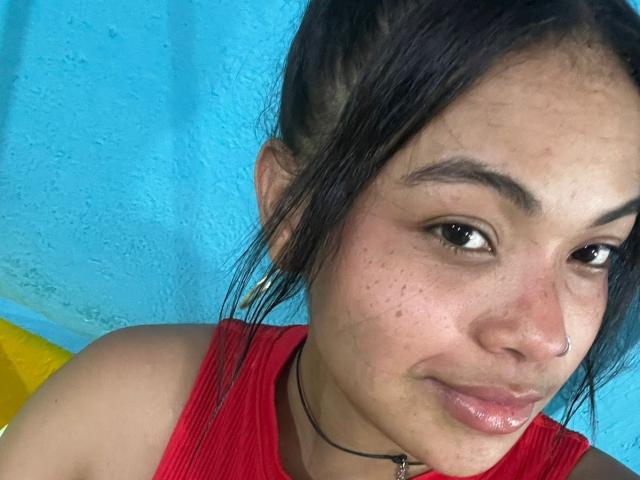 SoleneFontaine with a nose ring, dark hair in a messy bun, wearing a red top, black choker, and a slight smirk. She has small freckles and glossy lips.
