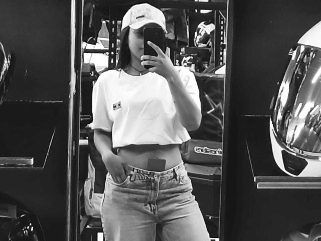 Black-and-white selfie of SoleneFontaine with medium tits, wearing a white crop top, blue jeans, and a baseball cap, standing in a cluttered room, holding a phone.