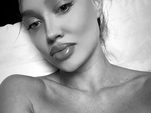 Black-and-white photo of BerryBoo with glossy lips, slightly parted, bare shoulders, and dark, wavy hair, looking seductively at the camera. Her face is smooth, with subtle makeup.