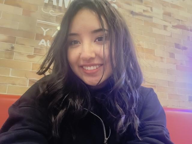 SusanMonroy with wavy black hair, smiling, wearing a black hoodie, sitting on a red bench, brick wall in the background, light skin, cute, slightly messy hair, happy expression.