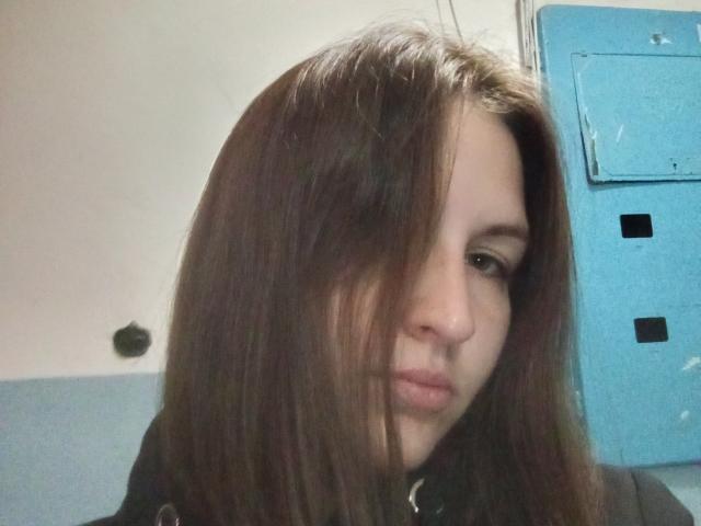 Vladlina has long, straight brown hair covering her right eye. She has a fair skin tone and slightly pursed lips. She wears a black top with a silver collar. The blue wall and electrical panel are in the background. Her expression is neutral. The image has a slightly blurry texture. The lighting is soft.