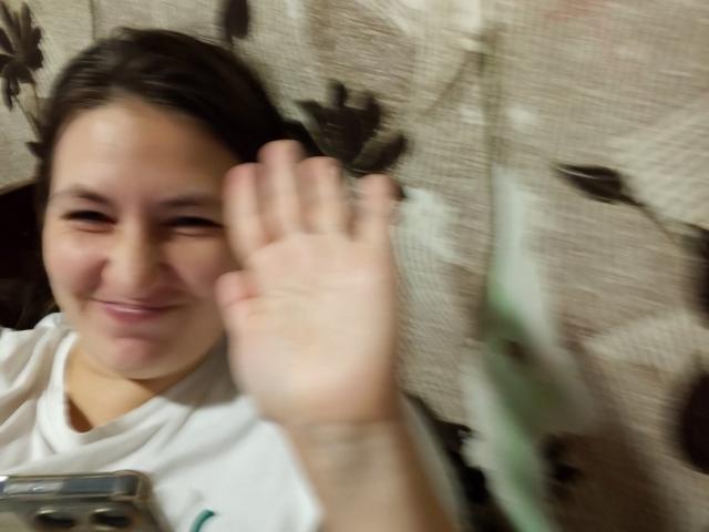 Close-up of Vladlina with a blurred background. Vladlina, a young woman with light skin and dark brown hair, is smiling slightly and waving with her right hand. She is wearing a white T-shirt with a green graphic on the left side. Her face is centered in the frame, with the blurred background showing a beige fabric with dark brown leaf patterns. The image has a soft, slightly out-of-focus quality. Her hand is in the foreground, partially obscuring the background.