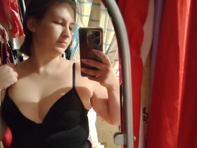 Vladlina takes a mirror selfie with a smartphone. She has a light skin tone, brown hair in a loose bun, and wears a black, low-cut, spaghetti-strap top that reveals her cleavage. Her right hand pulls at the top's strap, and her left hand holds the phone. The phone is dark with a visible camera on the back. The background has red and patterned curtains, with some clothing items hanging. The image is slightly blurry and has a casual, everyday feel.