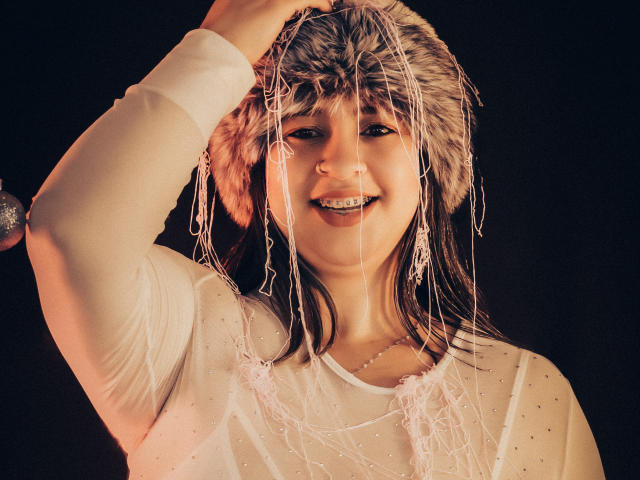 MelindaBoobs with a furry hat, smiling, wearing a white sheer top with sparkles, raising her arm, strands of white fabric hanging in front of her face.