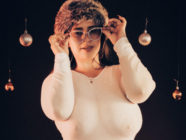 MelindaBoobs with big tits, wearing a furry hat, white long-sleeve shirt, and oversized glasses, adjusting the hat, with shiny ornaments hanging in the dark background.