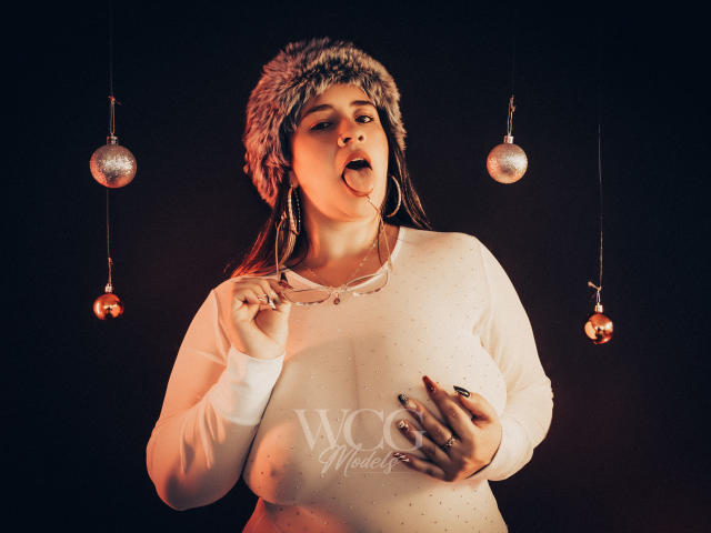 MelindaBoobs with tongue out, wearing a fur hat, white long-sleeve shirt, holding a candy cane, large tits, dark background, hanging ornaments, and "WMC Models" watermark.