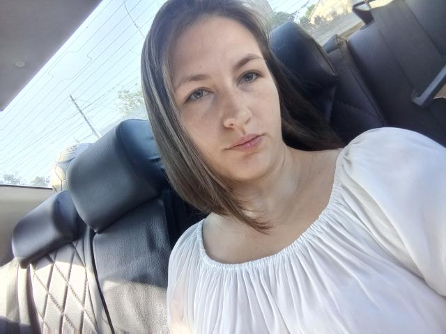 Vladlina is a young woman with fair skin and straight brown hair, sitting in a car. She wears a white, puffed-sleeve blouse with a gathered neckline. Her expression is neutral, and she looks directly at the camera. The car's black leather seat is visible behind her, with a window allowing sunlight to illuminate her face. The background shows blurred outdoor elements. Natural light highlights her clear skin and subtle makeup.