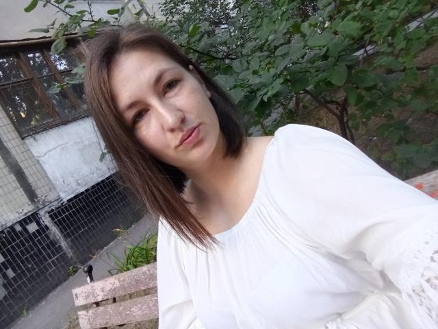 Vladlina has light skin, shoulder-length brown hair, and a neutral expression. She's wearing a white, loose-fitting, long-sleeve top. The image focuses on her upper body and face. She's slightly tilted to the right, with green leaves and a white building in the background. Natural light highlights her face, and the texture of her top is visible. The composition is casual and slightly angled.