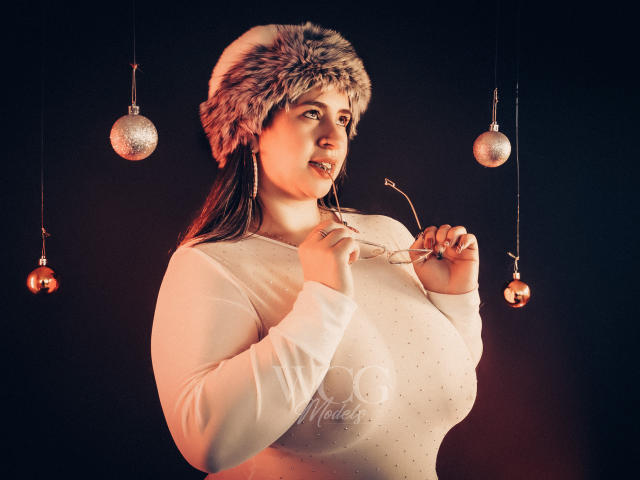 MelindaBoobs with large tits, wearing a white, long-sleeved, dotted top and fur hat, holding glasses, looking to the side, with two silver ornaments hanging in the background.