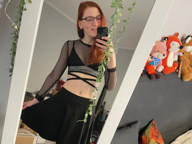 FinaFoxy-hot with red hair, glasses, black mesh top, strappy black skirt, holding phone, mirror selfie, green plant, stuffed animals on gray couch, smiling, showing belly button.
