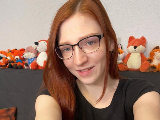 FinaFoxy-hot with red hair, glasses, and black shirt, smiling at camera. Stuffed foxes in background. Her tits are small, she's cute, and has a slight nose freckle.