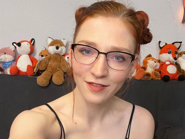 FinaFoxy-hot with red hair in buns, glasses, pale skin, and pink lips, wearing a black spaghetti strap top, surrounded by stuffed foxes.
