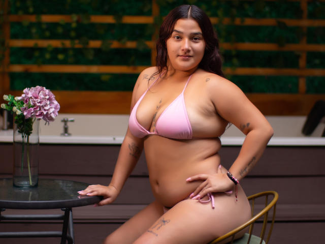 GabyCurvy with curvy, tan skin, large tits, and tattoos in a pink bikini, sitting at a table with a flower vase, hand on hip, smiling confidently.