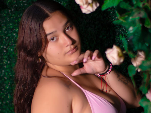 GabyCurvy with brown hair, wearing a pink halter top, looking at the camera, hand near chin, tattoo on right wrist, pink nail polish, rose in foreground.