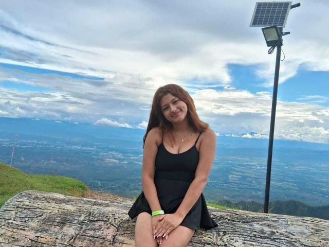 BrendaGreen, with long brown hair and black dress, sits on a rock, hands on knees, solar panel in background, blue sky and clouds above, smiling slightly.