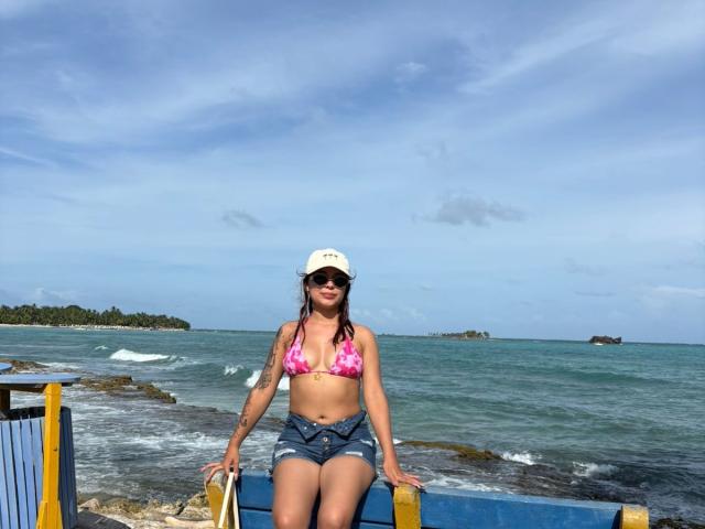 EileenSwift sits on a blue and yellow metal structure by the ocean. She wears a pink tie-dye bikini top, denim shorts, a white cap, and dark sunglasses. Her right arm has a tattoo. The sea is turquoise with small waves, and a distant island is on the right. The sky is clear with a few clouds. Her hair is dark and loose. The image is a casual, beachfront photograph.
