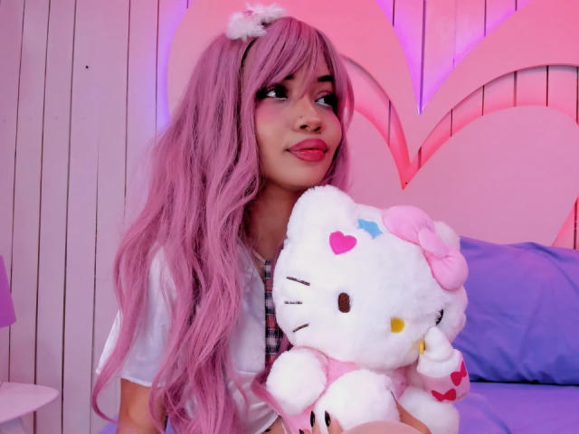 LiliMays with pink hair, wearing a white shirt, holding a Hello Kitty plushie, tongue slightly out, pink and purple lighting, fluffy white headband, cute and playful expression.