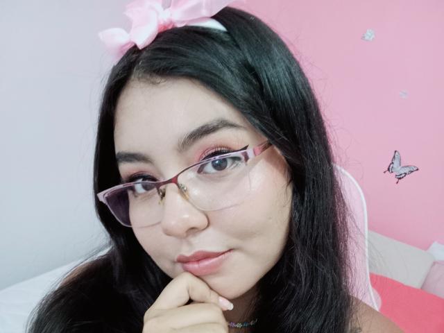 ValerieGreyX mit langem, schwarzem Haar, einem rosa Schleifchen, Brille und rosa Lidschatten, die sich ihr Kinn berührt, eine rosa Wand, ein weißes Bett mit rosa Details und ein Schmetterlingsaufkleber im Hintergrund..