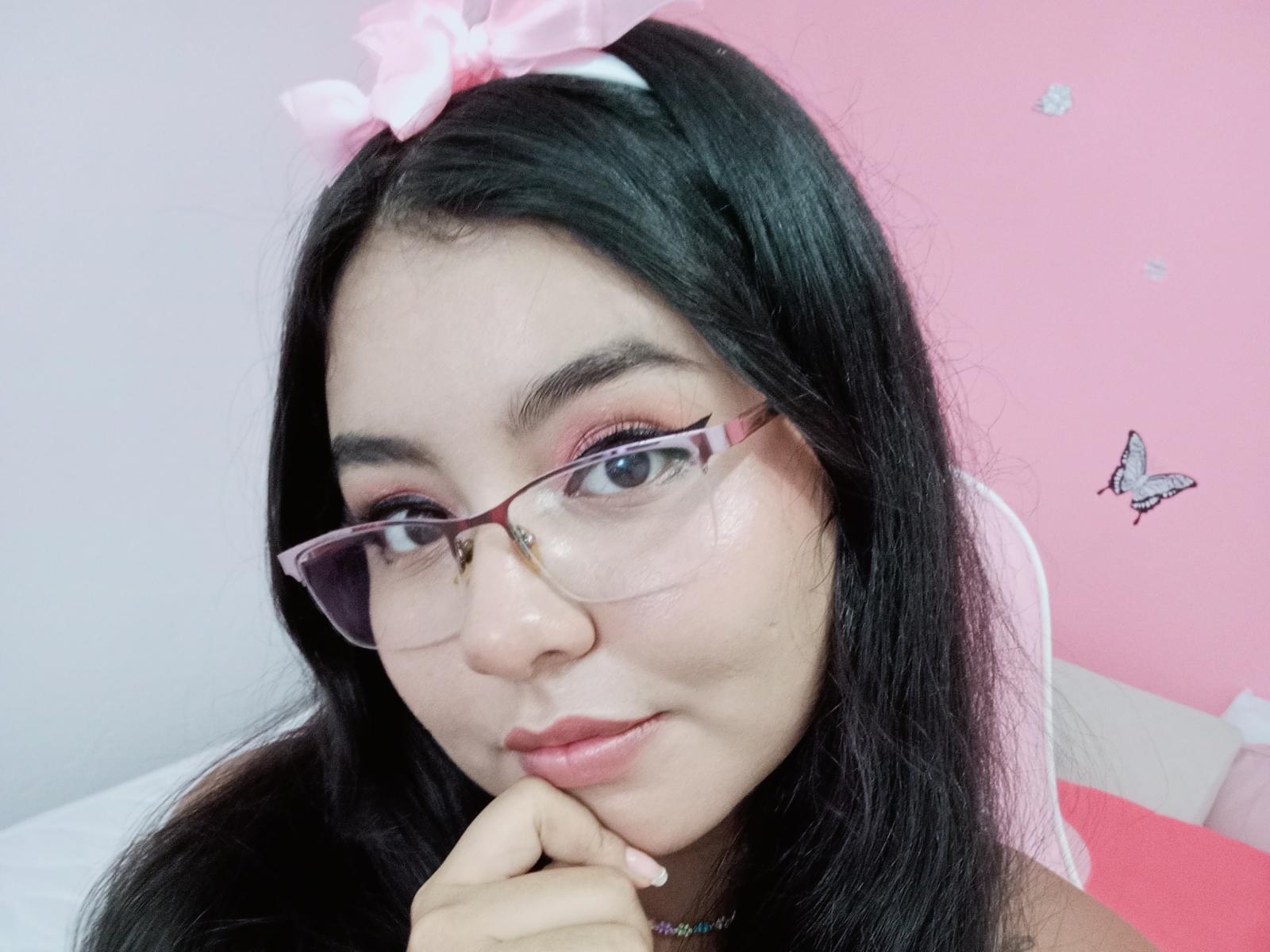 ValerieGreyX con cabello negro largo, un lazo rosa, gafas y sombra de ojos rosa, tocándose la barbilla, una pared rosa, una cama blanca y rosa, y una pegatina de mariposa en el fondo..