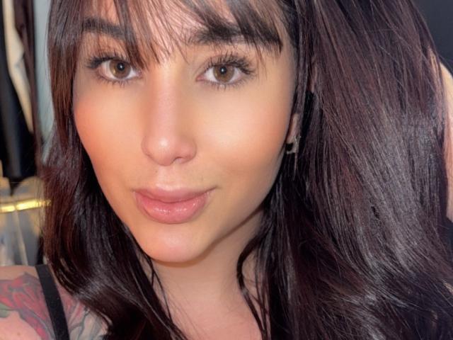 AgathaRuiz has brown eyes, full pink lips, and dark brown, wavy hair with bangs. She's making a pouty face, showing a tattoo on her right arm.