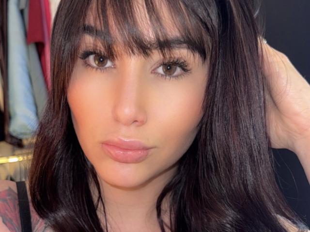 AgathaRuiz has dark brown eyes, full pink lips, and shiny black hair with bangs, looking serious with a hand in her hair, wearing a black top.